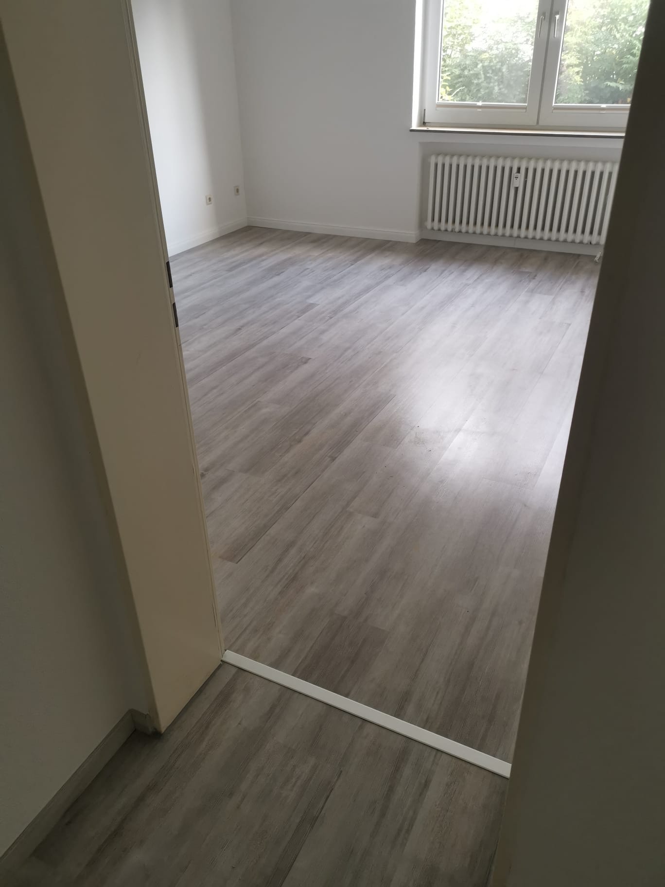 Ein Blick durch eine Türöffnung in einen hellen Raum mit einem neu verlegten, hellgrauen Laminatboden in Holzoptik. Im Hintergrund sind ein Fenster mit Heizkörper und weiße Wände sichtbar.