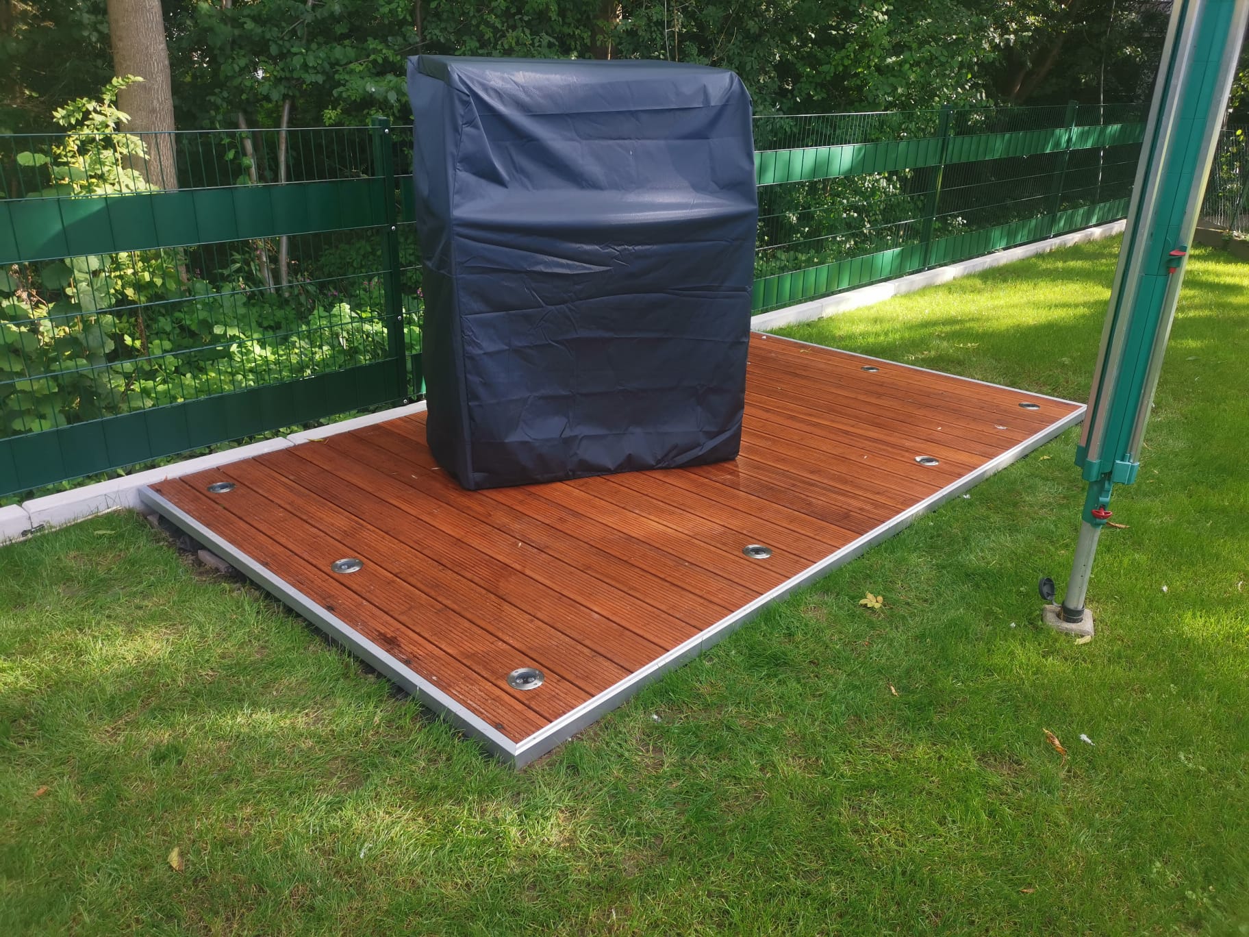 Aufnahme einer kleinen, quadratischen Terrasse aus braunen Holzdielen mit einer silberfarbenen Aluminiumumrandung, die auf einem gepflegten grünen Rasen liegt. In der Mitte der Terrasse steht eine rechteckige Sitzgruppe, die mit einer schwarzen, wasserabweisenden Plane abgedeckt ist. Im Hintergrund sind ein grüner Doppelstabmattenzaun und dichtes Grün von Bäumen und Büschen sichtbar.