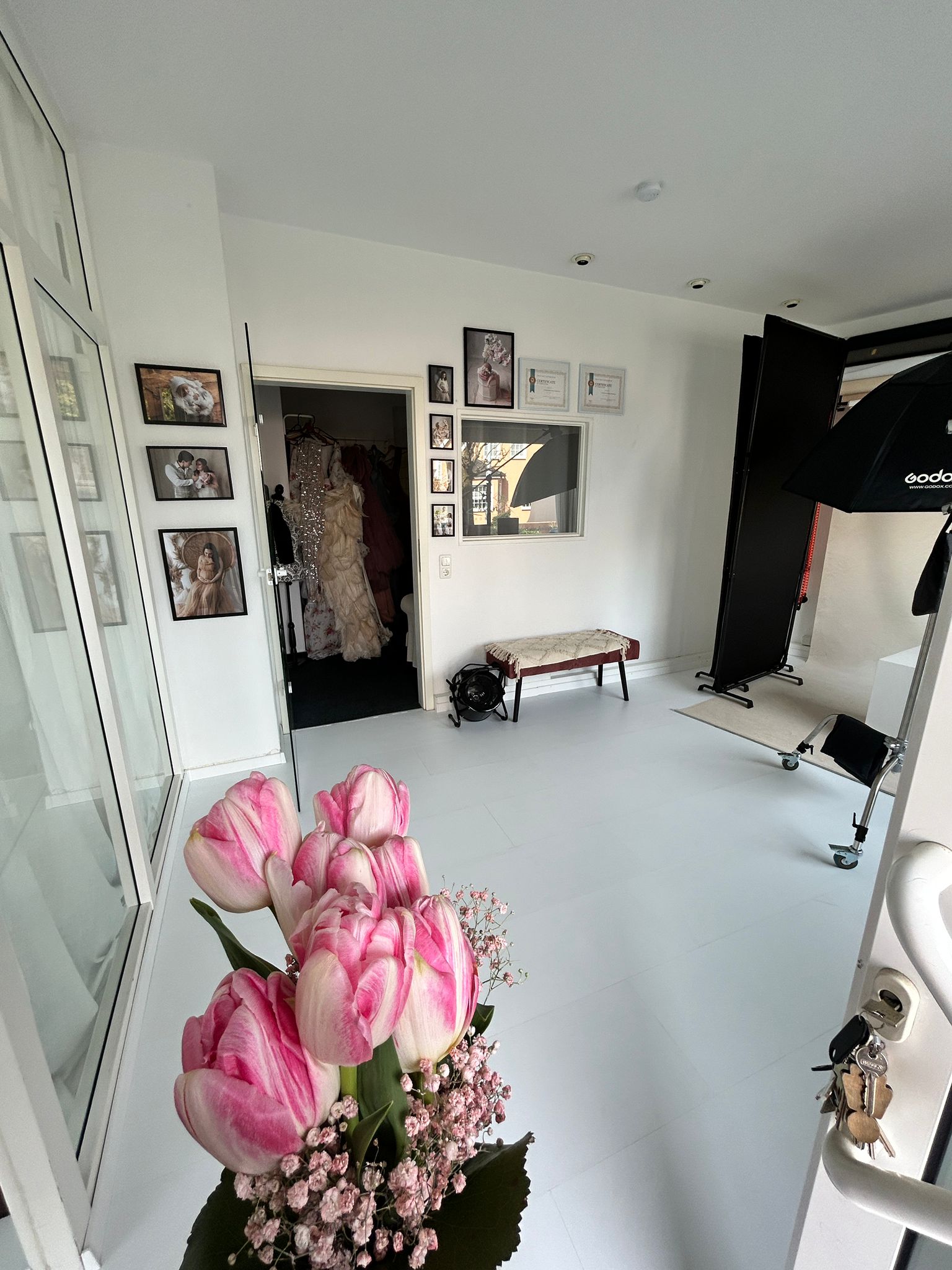 Ein Blick in einen hellen Raum mit weißem Boden. Im Vordergrund sind rosa-weiße Tulpen mit kleinen rosa Blüten zu sehen. Im Hintergrund, durch eine offene Tür, hängen mehrere aufwendige, helle Kleider. Im hinteren Teil des Raumes befindet sich Fotoequipment wie ein großer Softbox-Blitz.