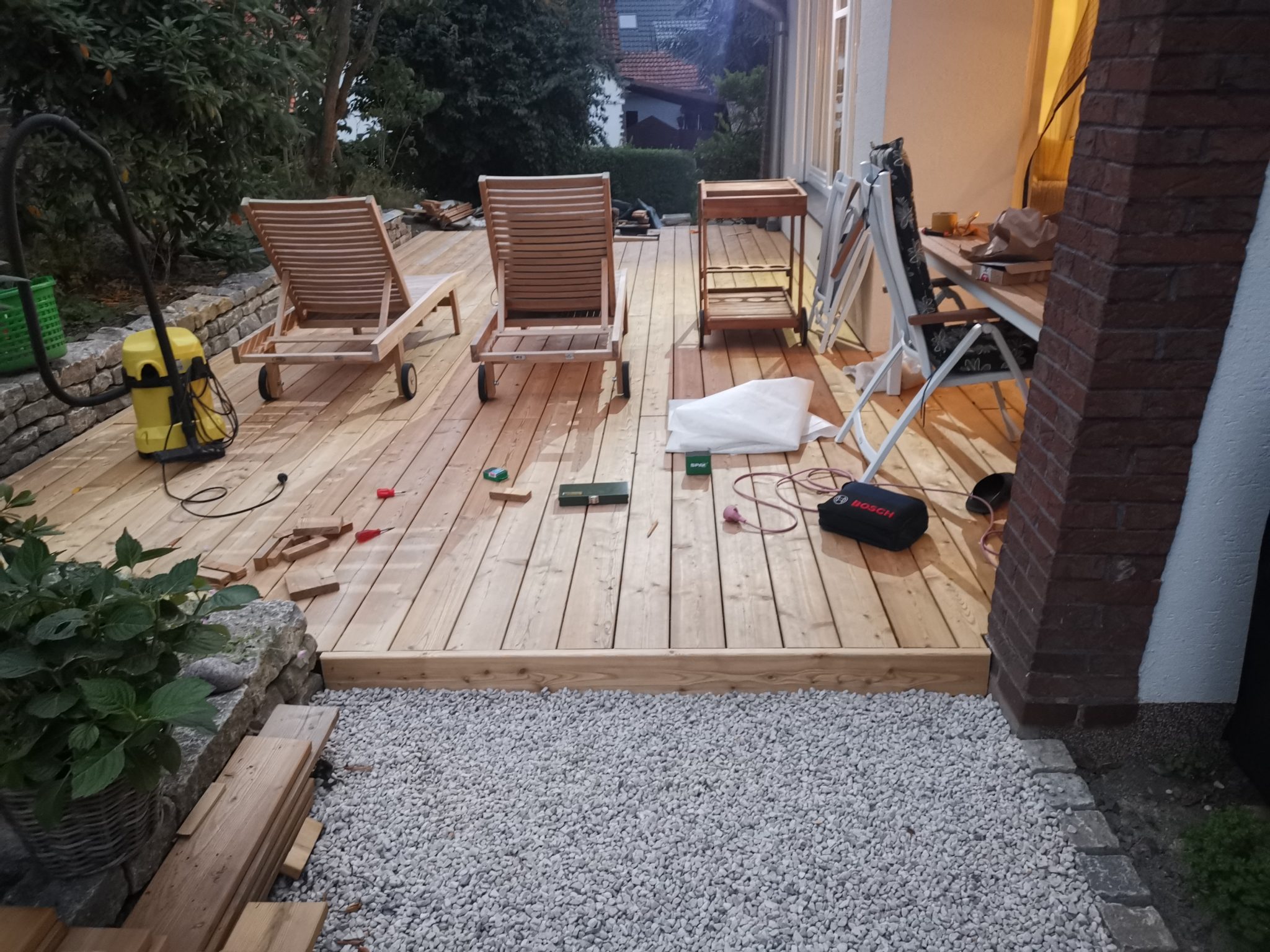Aufnahme einer Holzterrasse, bei der die hellen Holzdielen bereits verlegt sind. Verschiedene Werkzeuge wie ein Staubsauger, Kabel, eine Stichsäge und kleinere Handwerkzeuge liegen auf der Terrasse. Zwei hölzerne Liegestühle stehen ebenfalls darauf. Die Terrasse grenzt an ein Haus mit einer einzelnen Säule aus roten Backsteinen und einen Bereich, der mit hellem Schotter bedeckt ist.