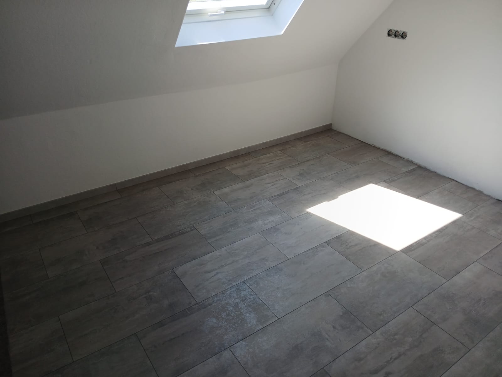 Ein Zimmer mit Dachschräge und einem neu verlegten, grauen Laminatboden in Fliesenbodenoptik ist zu sehen. Ein Dachfenster oben lässt helles Sonnenlicht in den Raum fallen.