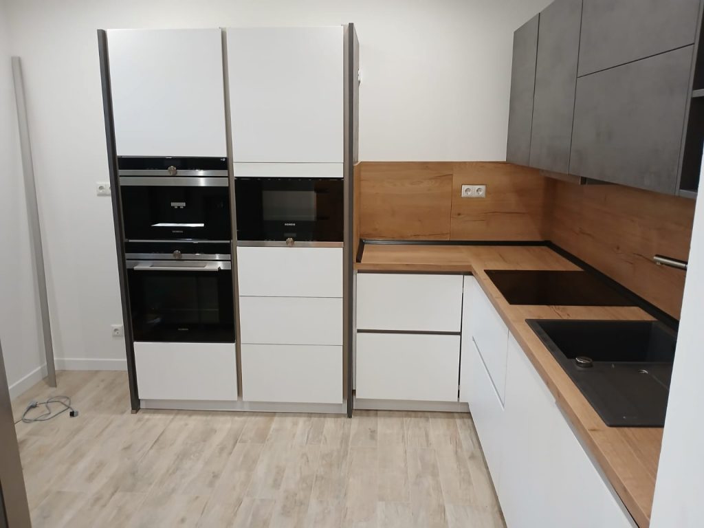 Aufnahme einer modernen Einbauküche mit weißen, grifflosen Unterschränken, einer Arbeitsplatte in heller Holzoptik mit integriertem Kochfeld und Spüle sowie grauen, schlichten Oberschränken. An der linken Seite befinden sich hoch eingebaute Geräte von Siemens (Backofen und Mikrowelle) und Neff (Kaffemaschine). Der Boden ist mit hellen, länglichen Holzdielen neu ausgelegt.