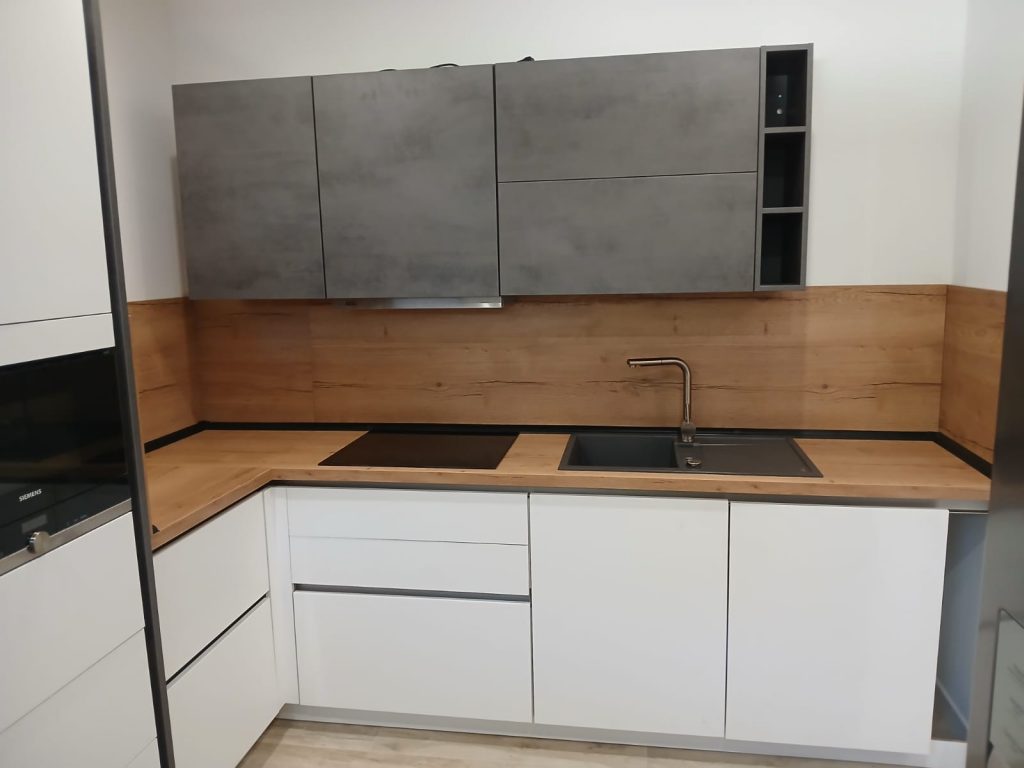 Aufnahme einer modernen Einbauküche mit weißen, grifflosen Unterschränken, einer Arbeitsplatte in heller Holzoptik mit integriertem Kochfeld und Spüle sowie grauen, schlichten Oberschränken. Der Boden ist mit hellen, länglichen Holzdielen neu ausgelegt.