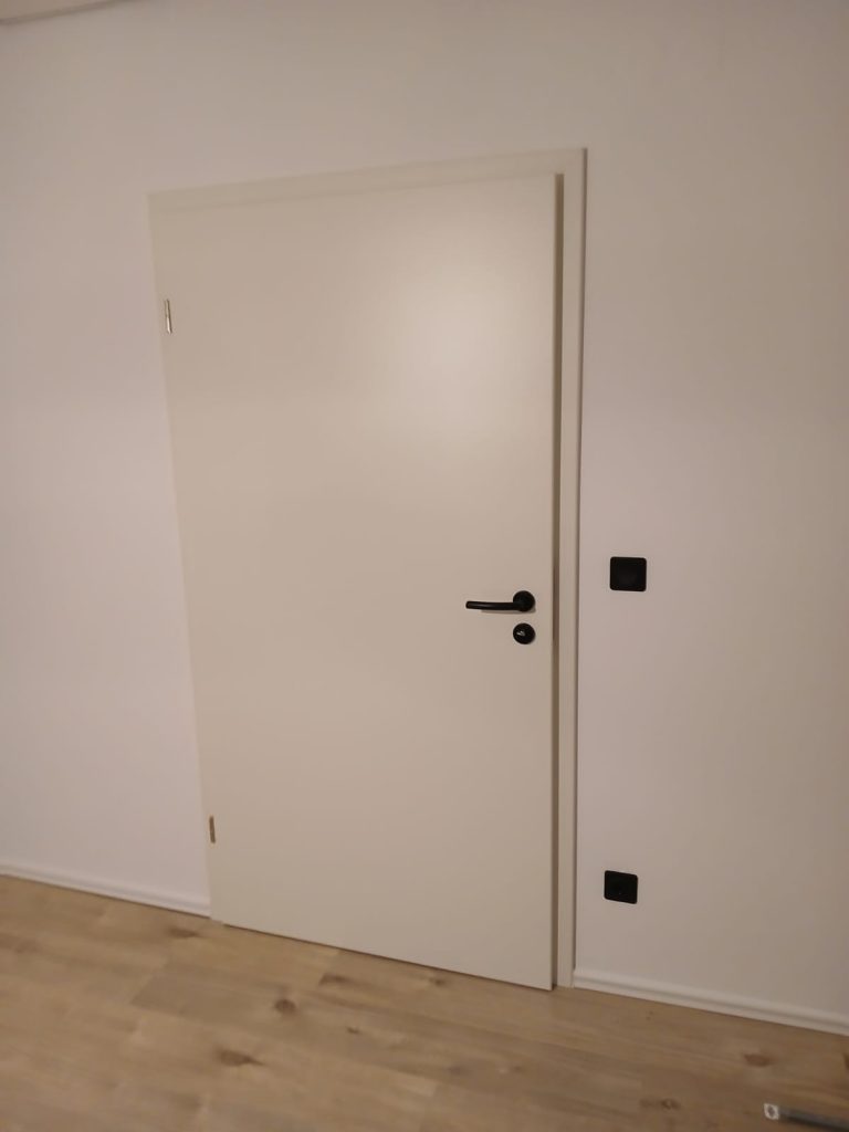 Eine neu eingebaute, weiße Innentür mit einem schwarzen Türgriff ist vor einer weißen Wand und einem hellen Holzboden zu sehen.