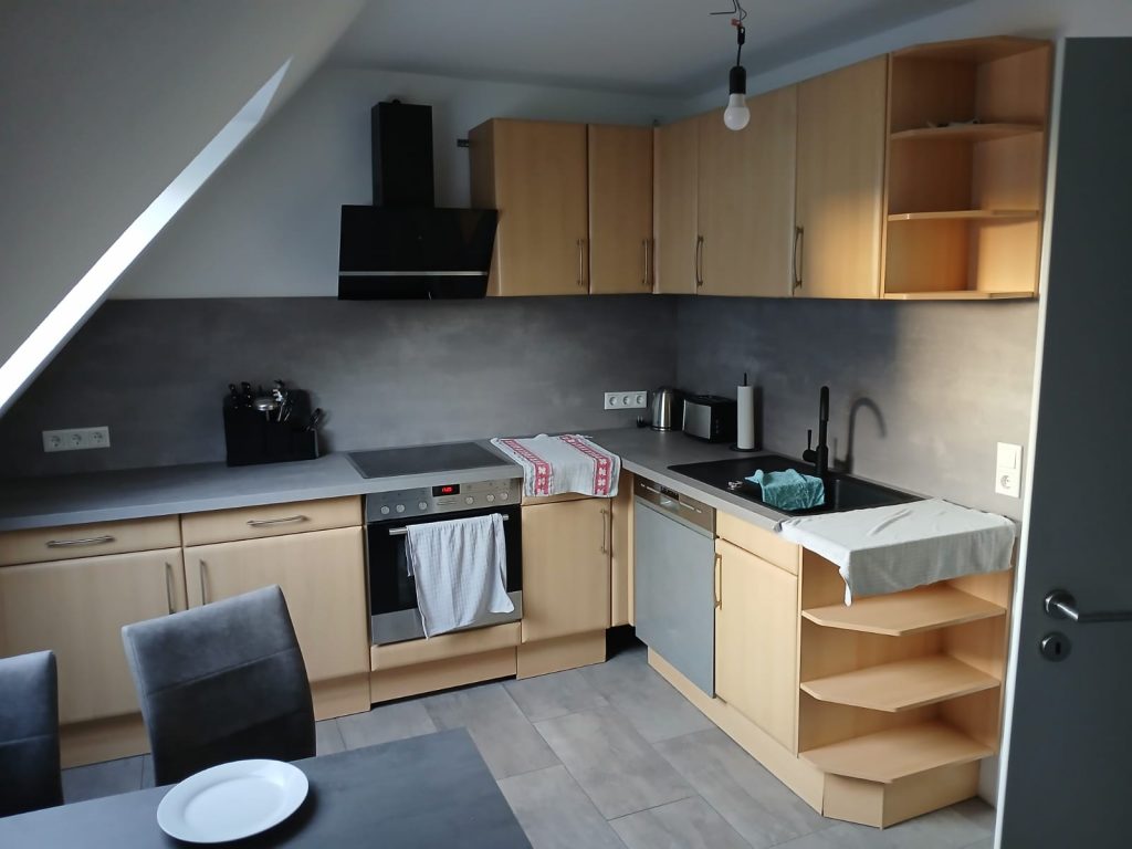 Aufnahme einer gut ausgestatteten Küche mit hellen Holzfronten, einer dunklen Dunstabzugshaube und einer grauen Arbeitsfläche mit Spüle und Herd. Der Boden ist mit hellgrauen, rechteckigen Fliesen neu ausgelegt. Ein grauer Esstisch mit zwei grauen Stühlen und weißen Tellern steht im Vordergrund.