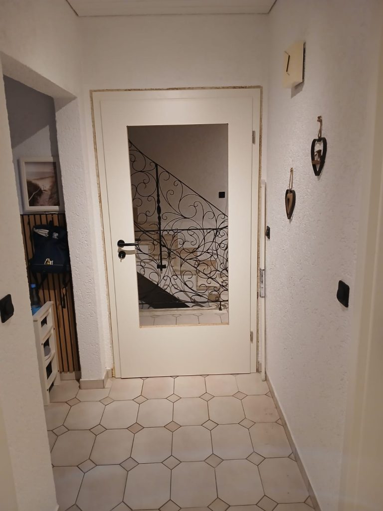Eine weiße Innentür mit einem rechteckigen Glaseinsatz ist in einem Flur mit hellen, gemusterten Fliesen zu sehen. Durch das Glas ist ein Treppenhaus mit einem schmiedeeisernen Geländer sichtbar.