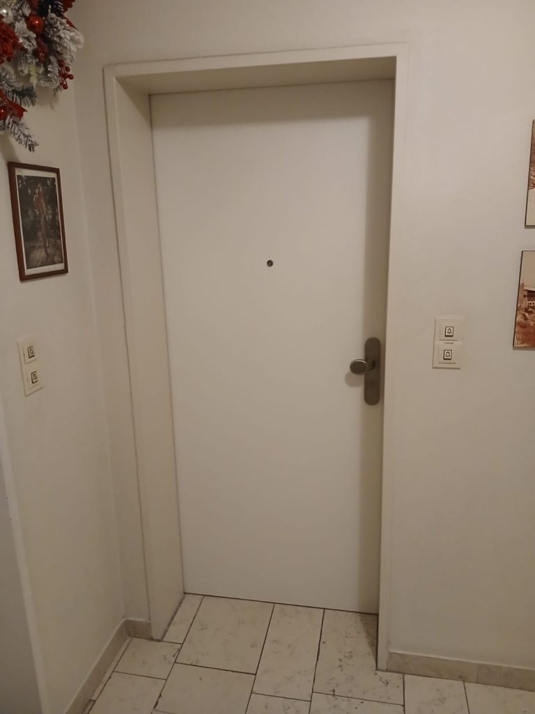 Eine geschlossene, weiße Wohnungseingangstür mit einem silberfarbenen Türgriff und einem Spion ist in einem Flur mit einem hellen, gemusterten Fliesenboden zu sehen. An den weißen Wänden befinden sich Lichtschalter und Dekoration.