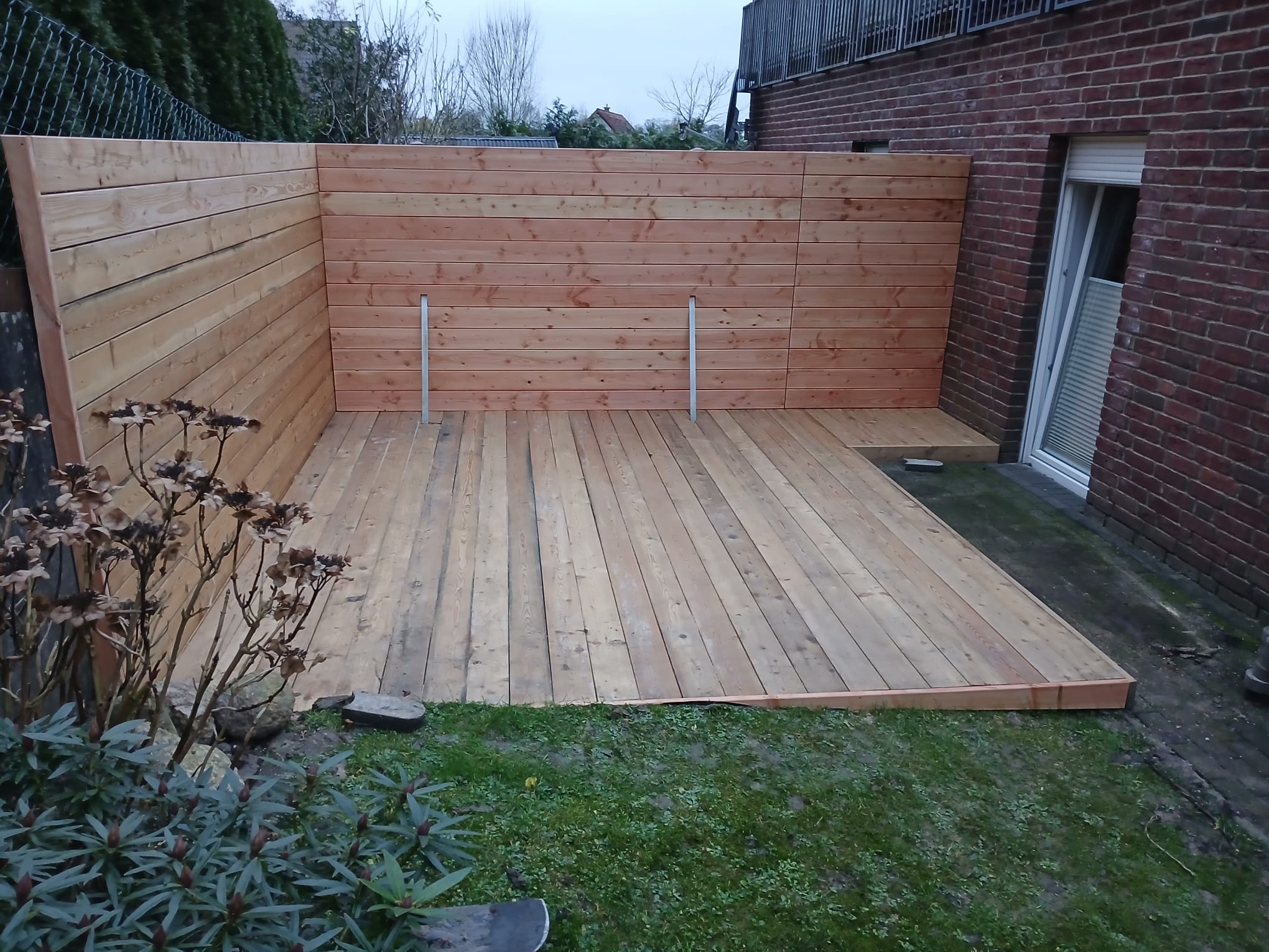 Aufnahme einer L-förmigen Holzterrasse aus hellen, behandelten Holzdielen, die direkt an die rote Backsteinwand eines Hauses anschließt. Die Terrasse ist von einem horizontal verlaufenden Holzzaun umgeben. Im Vordergrund ist ein Teil eines Gartens mit Rasen und Pflanzen sichtbar.