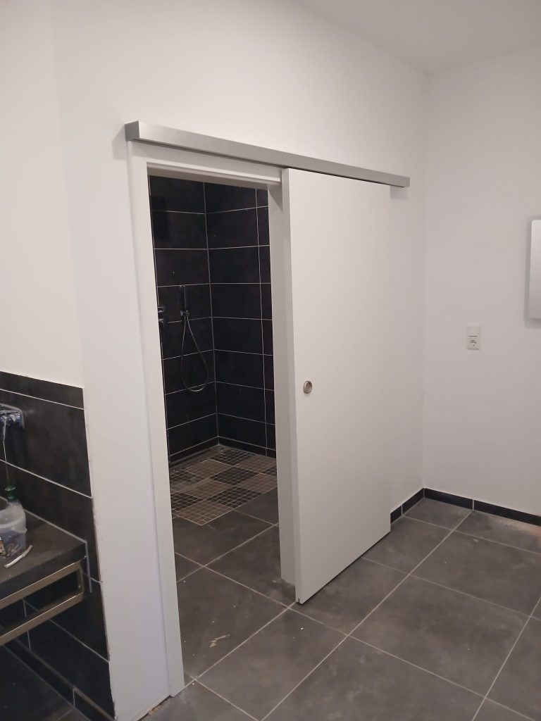 Eine teilweise geöffnete, weiße Schiebetür mit einer silberfarbenen Laufschiene ist vor einem Badezimmer mit dunklen Fliesen und einer Dusche zu sehen. Der Boden vor der Tür ist ebenfalls dunkel gefliest.
