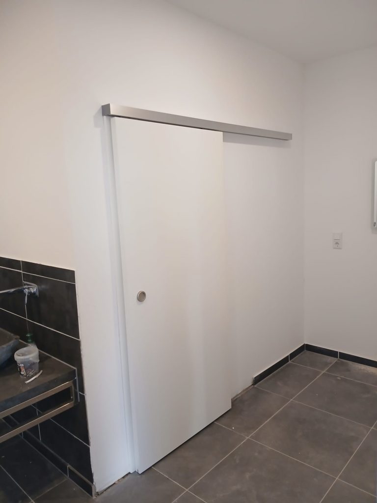 Eine geschlossene, weiße Schiebetür mit einer silberfarbenen Laufschiene ist vor einem Badezimmer mit dunklen Bodenfliesen zu sehen. Links ist ein Teil eines Waschtischs mit dunklen Fliesen sichtbar.