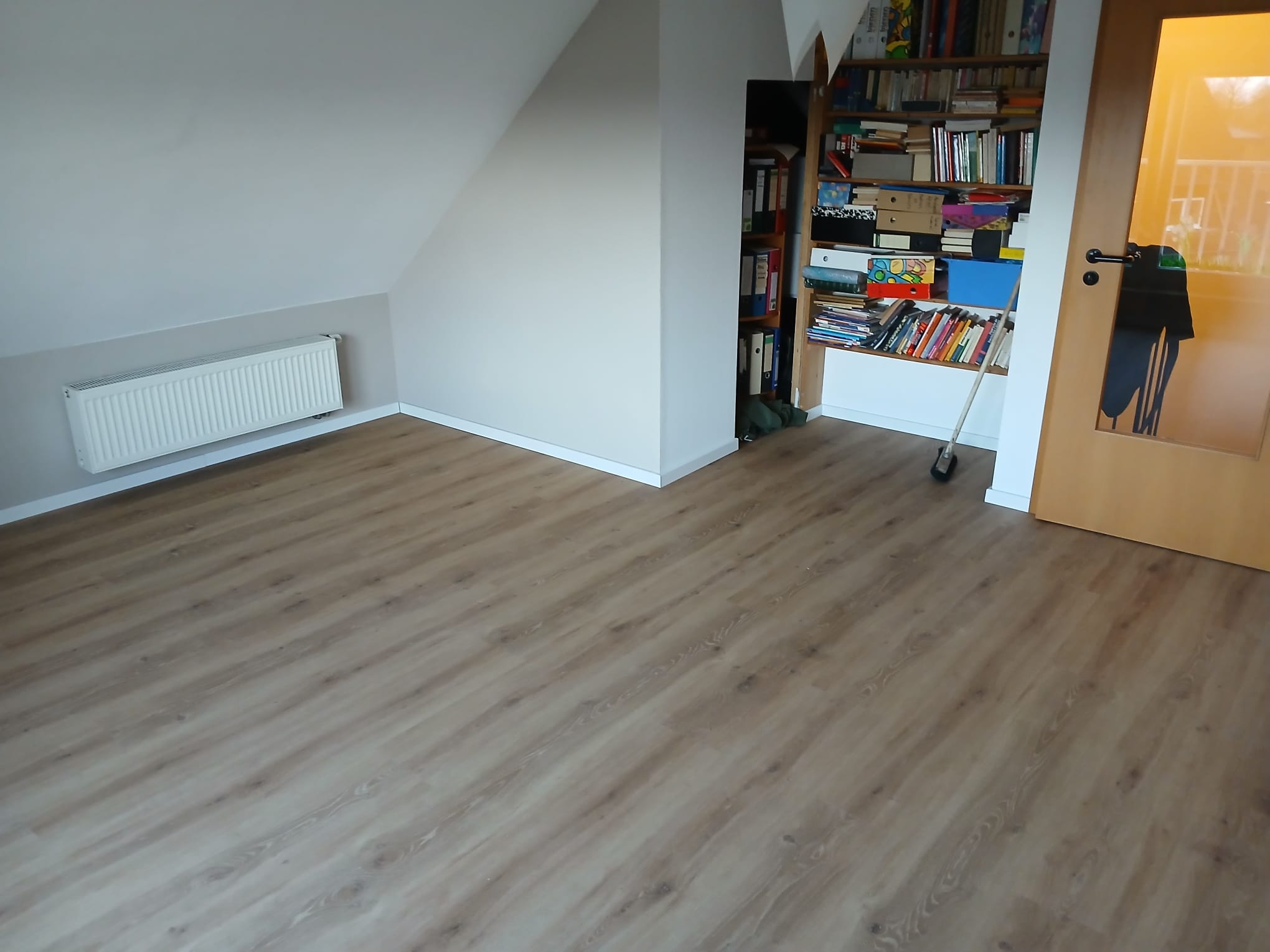 Ein Zimmer mit Dachschräge und einem neu verlegten, hellbraunen Holzboden ist zu sehen. An der linken Wand befindet sich ein Heizkörper, in der Ecke ein Bücherregal und rechts eine Holztür mit Glaseinsatz.