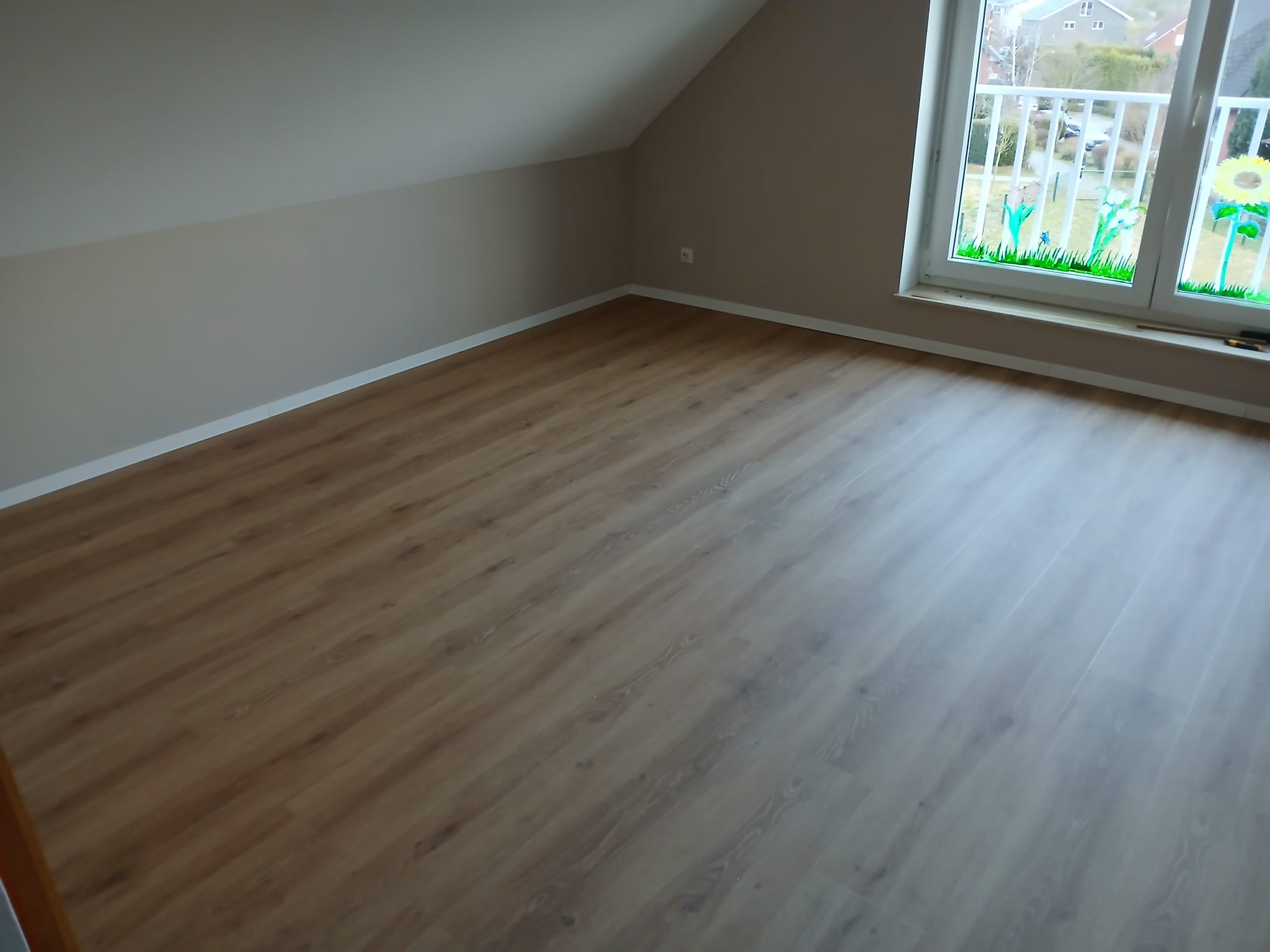 Frisch verlegter, heller Holzboden in einem hellen Dachgeschosszimmer mit einem Fenster, durch das Tageslicht einfällt.